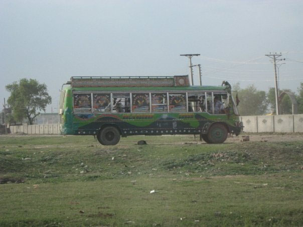  A green bus driving on a dusty road in a thrid world nation.